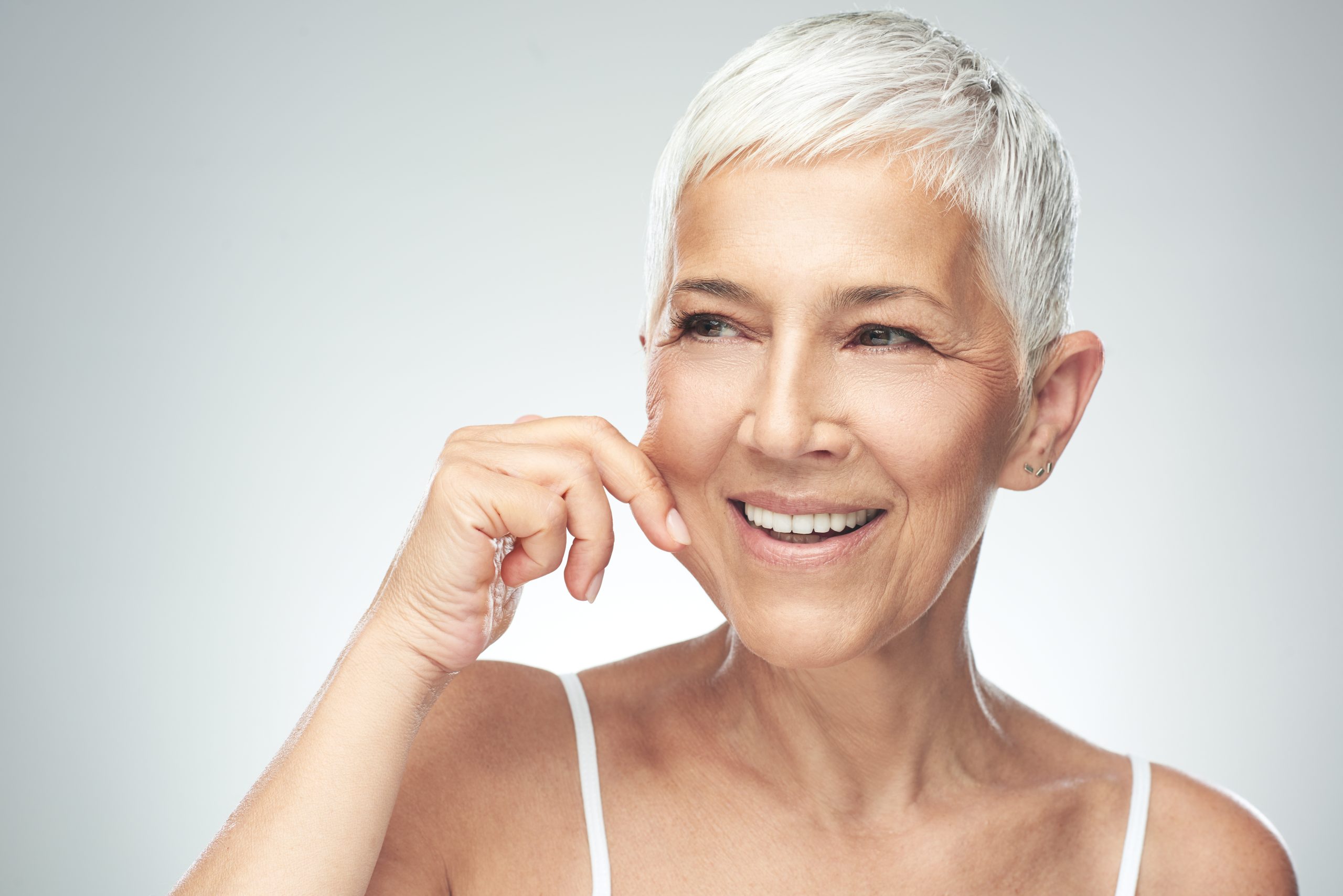 Gorgeous smiling Caucasian senior woman with short gray hair and natural beauty pinching her cheek. Beauty photography.